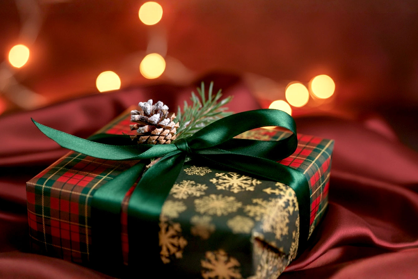 Professional studio photography, close-up shot of a beautifully wrapped Christmas gift using double-sided wrapping paper. One side of the paper features a classic red and green plaid, while a strategic middle fold reveals a contrasting, elegant gold snowflake pattern from the reverse side. The paper is crisp, with perfectly creased edges and an almost invisible seam. A simple, luxurious satin ribbon in deep forest green is tied neatly, adorned with a single, small, frosted pinecone and a delicate sprig of fresh holly. Soft, warm, diffused Christmas lights are subtly blurred in the background, creating a cozy, magical holiday ambiance. The focus is sharp on the gift wrap details, highlighting the texture and dual patterns. High dynamic range, rich colors, shallow depth of field.