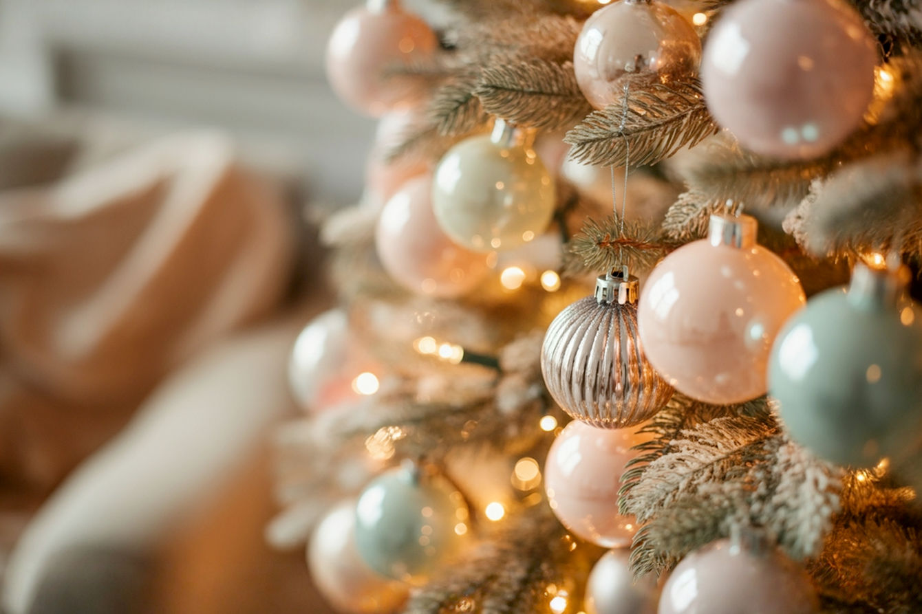 A close-up, high-angle professional photograph of a beautifully decorated Christmas tree adorned with pastel-colored ornaments and warm, soft lights. The ornaments should feature a mix of matte blush pinks, iridescent mint greens, subtle lavender, and pale gold metallics, catching the light elegantly. The background should be softly blurred, showing hints of a cozy, elegant living room setting with diffused natural light, emphasizing a serene and sophisticated holiday atmosphere. Lifelike, pro photography style.