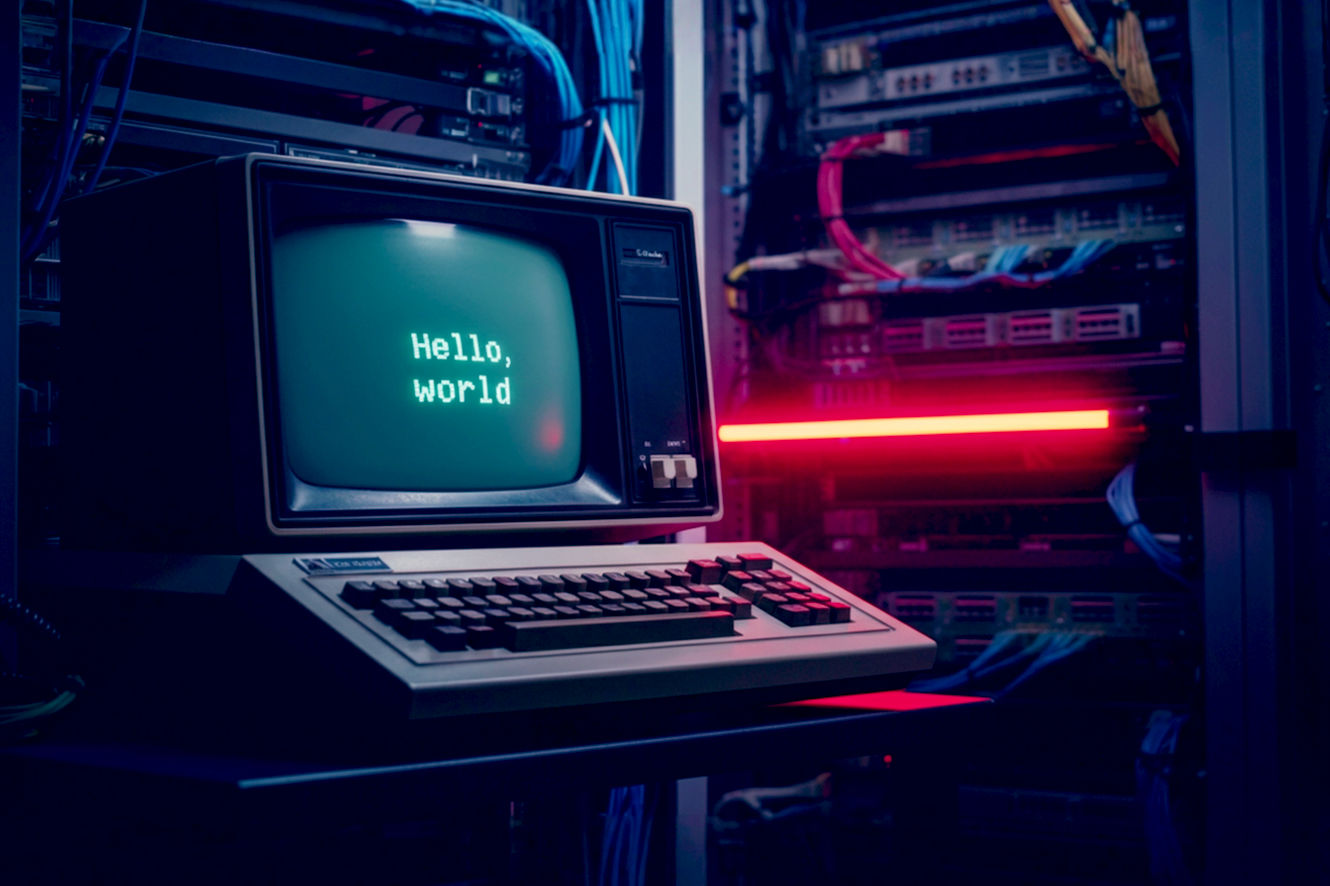 National Geographic style photograph of a vintage 1970s computer terminal (like a DEC VT100) alone in a dimly lit, server-filled room. The screen is the only bright light source, glowing with a green phosphor luminescence, displaying the iconic text: hello, world. The composition is tight on the terminal, with the background of server racks and cables softly out of focus. The lighting is dramatic and moody, with the single glow from the screen illuminating the dust motes in the air. The color palette is dominated by dark shadows, metallic grays, and the piercing green of the text. The mood is one of quiet, profound significance—the moment a machine first speaks.
