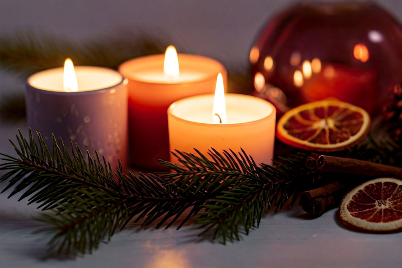 Professional, lifelike photograph of a collection of Christmas scented candles. The composition is a close-up, shallow depth of field shot, focusing on 3-4 elegantly designed candles, with at least two actively burning, casting a soft, warm glow. Interspersed among the candles are natural festive elements: fresh green pine sprigs, whole cinnamon sticks, and dried orange slices. The background is softly blurred, hinting at a cozy home interior with subtle, out-of-focus fairy lights. Lighting is warm, inviting, and atmospheric, emphasizing the candle flames' gentle radiance. The mood is tranquil, luxurious, and deeply festive. High resolution, rich textures, pro photography style.