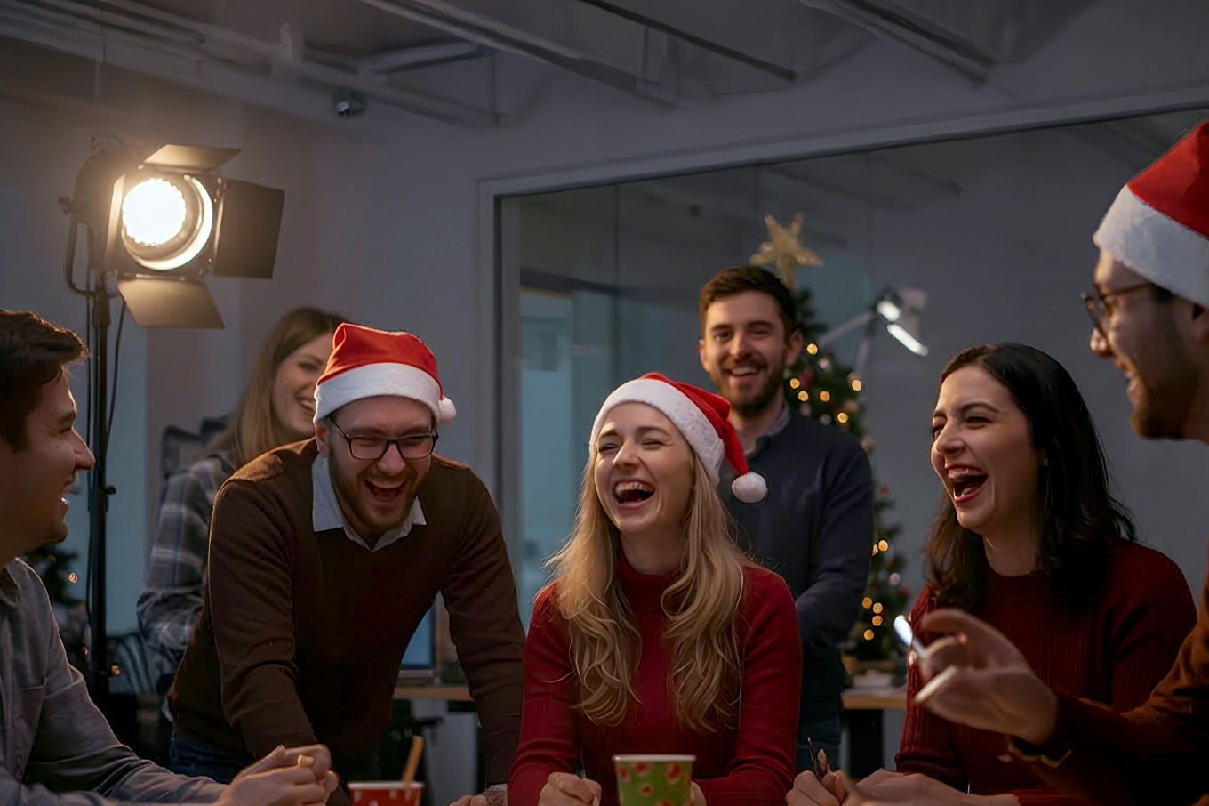 A vibrant, lifelike pro photography style image of a group of colleagues (mix of ages, genders) actively participating in a festive team building activity in a modern, well-lit office space. They are laughing and interacting positively. The scene should convey a sense of joy, collaboration, and holiday spirit. Elements could include subtle Christmas decorations in the background, perhaps a festive snack table, or a shared project they are working on together. The lighting should be warm and inviting, capturing candid, authentic moments of connection. The focus should be on the people and their positive interactions.