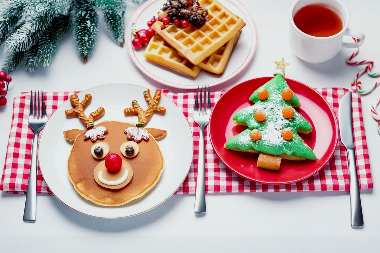 A top-down, lifelike professional photograph of a beautifully set Christmas breakfast table. The table is adorned with a festive red and white checkered tablecloth. In the center, there's a plate of perfectly round, fluffy pancakes artfully decorated to resemble cheerful reindeer, complete with pretzel antlers, mini chocolate chip eyes, and a bright red candy nose. Beside it, a stack of golden-brown waffles is transformed into a vibrant green Christmas tree, lightly dusted with powdered sugar and adorned with colorful sprinkles and small fruit pieces.