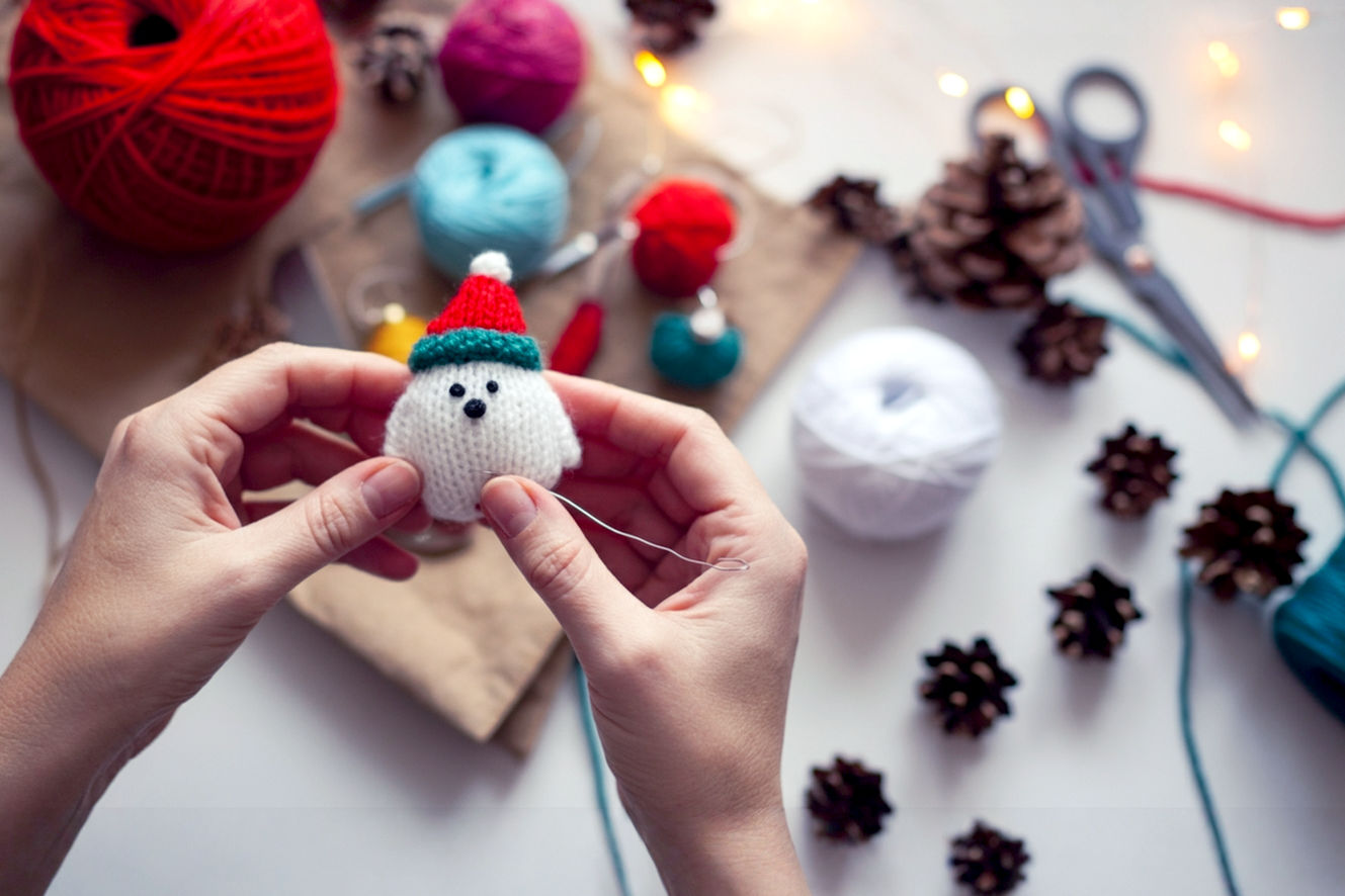 Pro photography style, lifelike, high-resolution image of a cozy, well-lit crafting scene. The main subject is a pair of hands (adult or older teen) actively engaged in making a 'Rustic Pinecone Ornament' or a 'Felt Christmas Tree Ornament'. The composition should be a close-up, slightly overhead shot focusing on the hands and the craft in progress, with a blurred background showing other festive, unfinished craft supplies like colorful felt, glitter, or small pinecones, and a hint of warm, twinkling Christmas lights. The lighting is soft, warm, and inviting, suggesting a cozy indoor setting. The mood is joyful, creative, and relaxed, emphasizing the 'easy DIY' aspect. Core theme: accessible, fun, and beautiful homemade Christmas crafting.