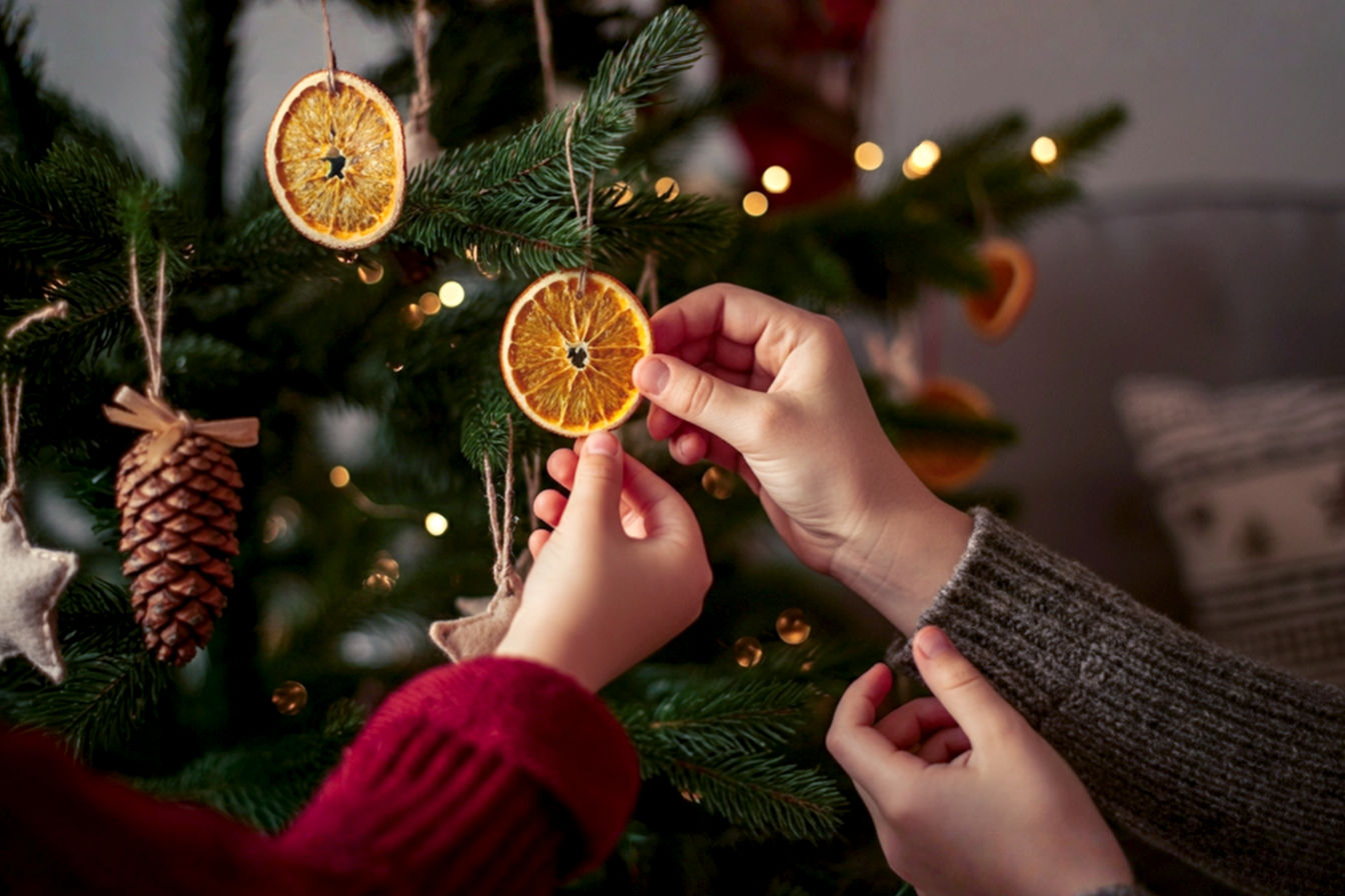 A warm, inviting, professional photography style image of a cozy living room during the Christmas season. The focus is on a beautifully decorated Christmas tree adorned with a mix of homemade, rustic-chic DIY ornaments, such as dried orange slices, salt dough stars, and pinecone accents. In the foreground, hands (one adult, one child) are gently placing a simple, charming felt ornament on a branch. Soft, golden fairy lights glow from the tree and a nearby mantelpiece, which is decorated with a DIY garland of cranberries and popcorn. A blurred background shows a comfortable sofa with a handmade Christmas pillow. The lighting is soft, warm, and golden, creating a cozy and nostalgic mood. The composition is a close-up, shallow depth of field shot, emphasizing the handmade details and the interaction. Lifelike, high-resolution, professional photography.