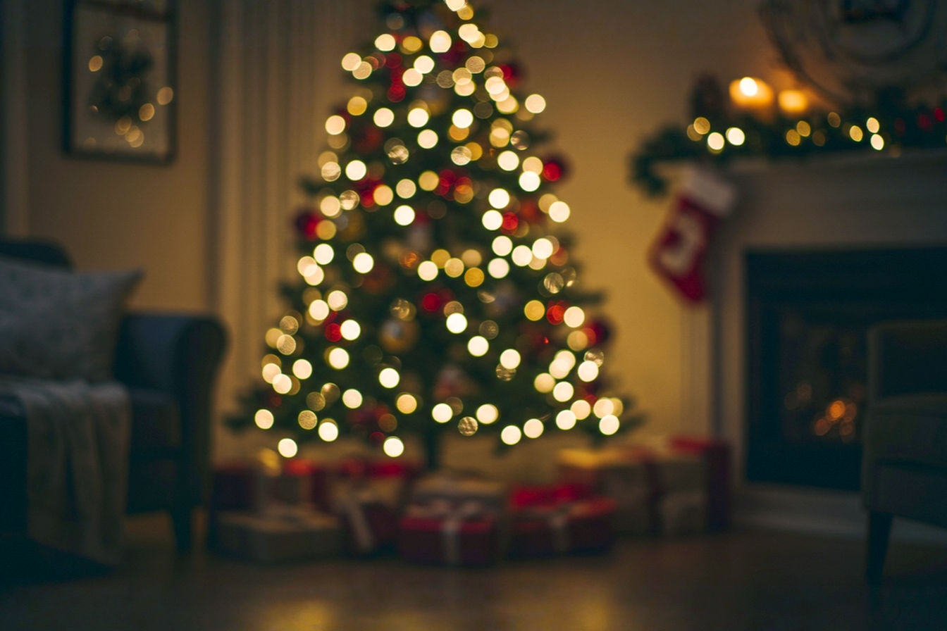 Professional, lifelike photograph of a beautifully decorated artificial Christmas tree standing elegantly in a cozy, modern living room. The tree is pre-lit with warm white LED lights, showcasing realistic, full branches with a mix of PE and PVC needles, adorned with classic red and gold ornaments. A soft, inviting glow emanates from the tree, casting gentle light on a festive scene. In the foreground, a few carefully wrapped presents are visible. The background features a blurred, warm interior with subtle holiday decor, suggesting comfort and joy. The composition is eye-level, capturing the tree's grandeur. Lighting is soft, warm, and inviting, emphasizing the tree's beauty and the room's festive ambiance. The mood is peaceful, joyful, and sophisticated. Focus on sharp detail of the tree and ornaments, with a shallow depth of field to highlight the main subject.