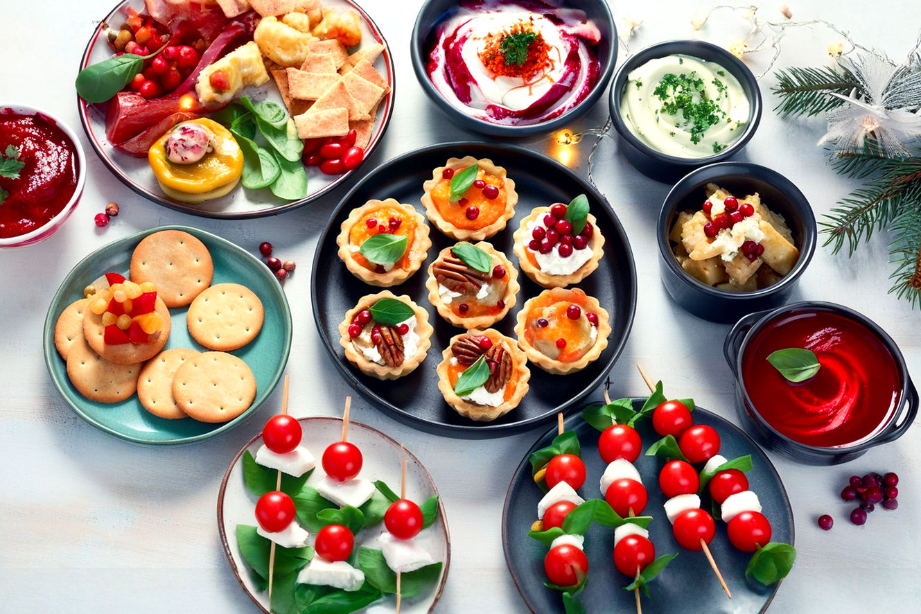 A vibrant, top-down, lifelike professional photography shot of a beautifully arranged holiday appetizer spread on a rustic wooden table. The scene features an assortment of colorful, bite-sized finger foods, including mini quiches, cranberry-pecan goat cheese truffles, caprese skewers, and various festive dips with crackers. Soft, warm ambient lighting illuminates the food, creating a cozy and inviting atmosphere. There are hints of holiday decor like subtle fairy lights or pine sprigs in the blurred background. The composition is abundant and artfully arranged, emphasizing the "hands-on" and "holiday" theme. The image should evoke a sense of warmth, deliciousness, and festive celebration.