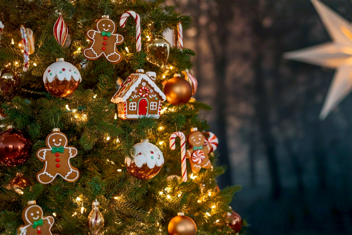 A professional, lifelike photograph of a beautifully decorated gingerbread themed Christmas tree. The tree is full, adorned with warm white lights, a mix of realistic-looking gingerbread men, gingerbread houses, and candy cane ornaments. The color palette is dominated by rich browns, creamy whites, and festive reds, with subtle gold accents. The tree is set in a cozy, softly lit living room, with a blurred background suggesting a warm, inviting home environment. There are a few wrapped gifts with coordinating paper and bows at the base. The lighting is soft and warm, highlighting the textures of the ornaments and creating a magical, nostalgic mood. Focus on the intricate details of the gingerbread decorations, making them appear almost edible.