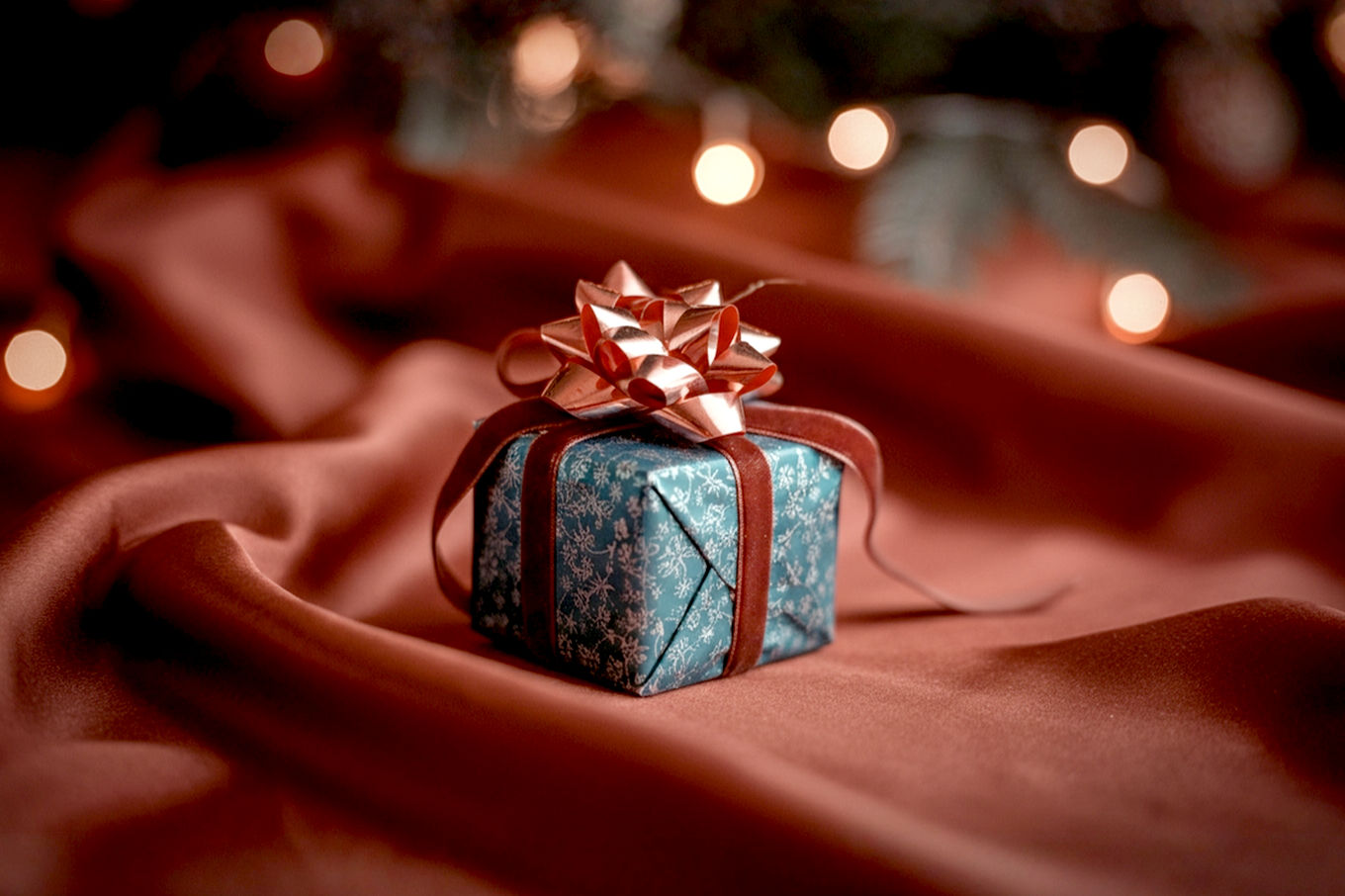Lifelike professional photography of a beautifully wrapped small Christmas gift box, perhaps nestled in a cozy setting with subtle holiday lights or a hint of a Christmas tree in the soft-focus background. The gift should be elegant and intriguing, hinting at a thoughtful surprise within, rather than a specific item. Emphasize warm, inviting lighting, rich textures (like velvet ribbon or textured paper), and a sense of coziness and anticipation. The focus should be on the "small package" feeling special and impactful.