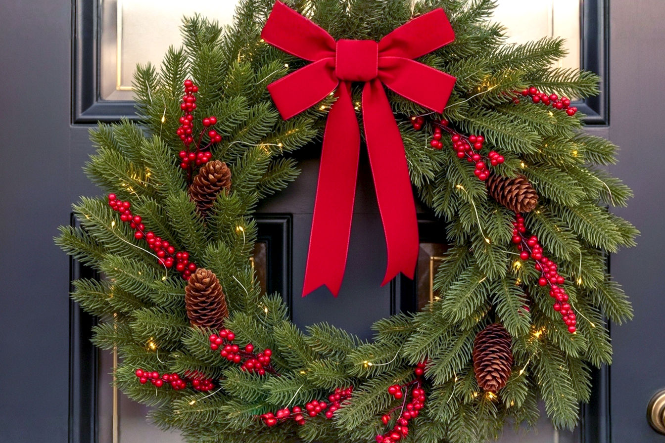 A professional, lifelike, high-resolution photograph of a beautifully decorated traditional Christmas wreath hanging on a dark, elegant front door. The wreath is lush with vibrant green fir branches, adorned with classic deep red berries, natural brown pinecones, and a rich, deep red velvet bow. The lighting is soft, warm, and inviting, suggesting late afternoon or early evening, with subtle, warm white LED fairy lights woven into the wreath emitting a gentle, magical glow. The composition is a close-up, slightly off-center shot, focusing on the intricate details and textures of the wreath and the elegant grain of the door. The background is softly blurred to emphasize the wreath. The mood is cozy, festive, and welcoming. This is a pro photography style image, highly realistic, with sharp focus on the wreath.