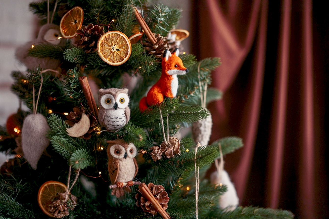 A lifelike, professional photograph of a beautifully decorated Christmas tree with a prominent woodland theme. The tree should be adorned with natural elements like real pinecones, dried orange slices, cinnamon sticks, and subtle wooden ornaments. Incorporate delicate, warm white fairy lights that cast a soft glow. Include a few tasteful, realistic-looking felt or wooden woodland animal ornaments (e.g., a small owl or fox peeking from branches). The composition should be warm and inviting, with a shallow depth of field to keep the focus on the decorations. Lighting should be soft and diffused, mimicking natural indoor light or early evening glow. The background should be slightly blurred, suggesting a cozy living room setting with hints of rustic textures like a fireplace or a knitted throw. The overall mood should be serene, enchanting, and authentic, emphasizing the natural charm.