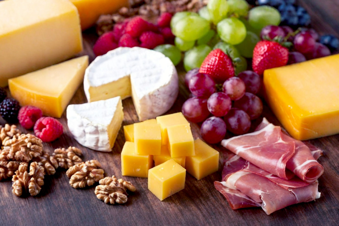 Lifelike professional photography of a lavish, overflowing Christmas charcuterie board. The board is made of dark, rustic wood and is centrally composed, filled with an abundance of festive, colorful ingredients. Prominently feature a variety of cheeses (creamy brie, aged cheddar, blue cheese), artfully folded cured meats (prosciutto, salami roses), and vibrant fresh fruits like red cranberries, green grapes, and sliced oranges. Include sprigs of fresh rosemary and sugared cranberries for a strong holiday feel. The lighting is warm and inviting, with soft, golden hues casting gentle shadows, creating a cozy and luxurious mood. The background is softly blurred, hinting at a festive holiday gathering, possibly with subtle fairy lights. The image should be sharp, high-resolution, and visually rich, emphasizing texture and color to evoke a sense of delicious indulgence and holiday cheer.