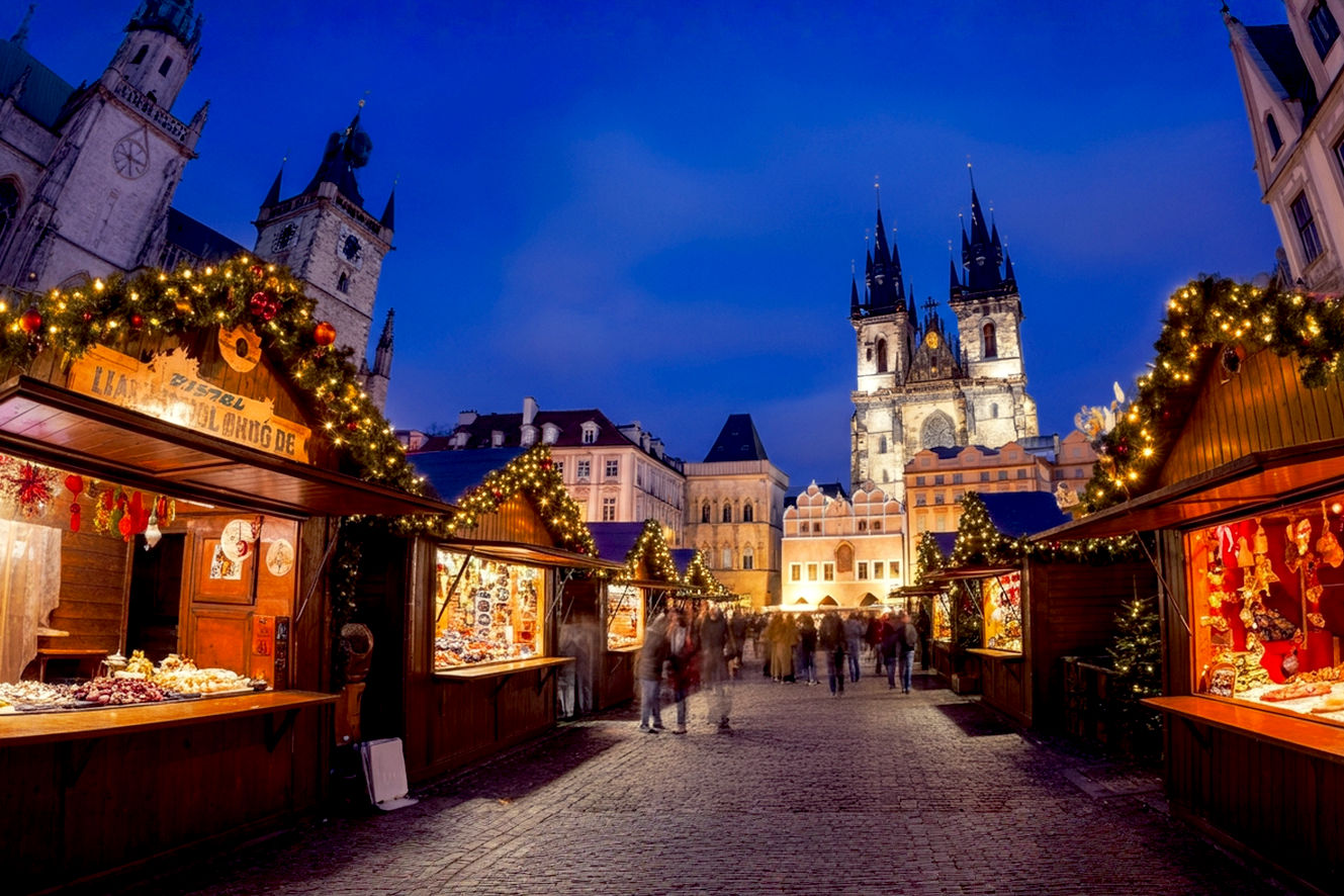 professional travel photography style image showcasing a magical Christmas destination scene. The composition should feature a European Christmas market at golden hour with warm, glowing vendor stalls displaying traditional holiday crafts and foods. In the foreground, show beautifully decorated wooden market stalls with twinkling fairy lights, steaming mugs of mulled wine, and colorful Christmas ornaments. The middle ground should include families and couples browsing the market in winter coats, creating a sense of warmth and community. The background should feature stunning Gothic or medieval architecture - cathedral spires or historic buildings - silhouetted against a twilight sky with the first stars appearing.
