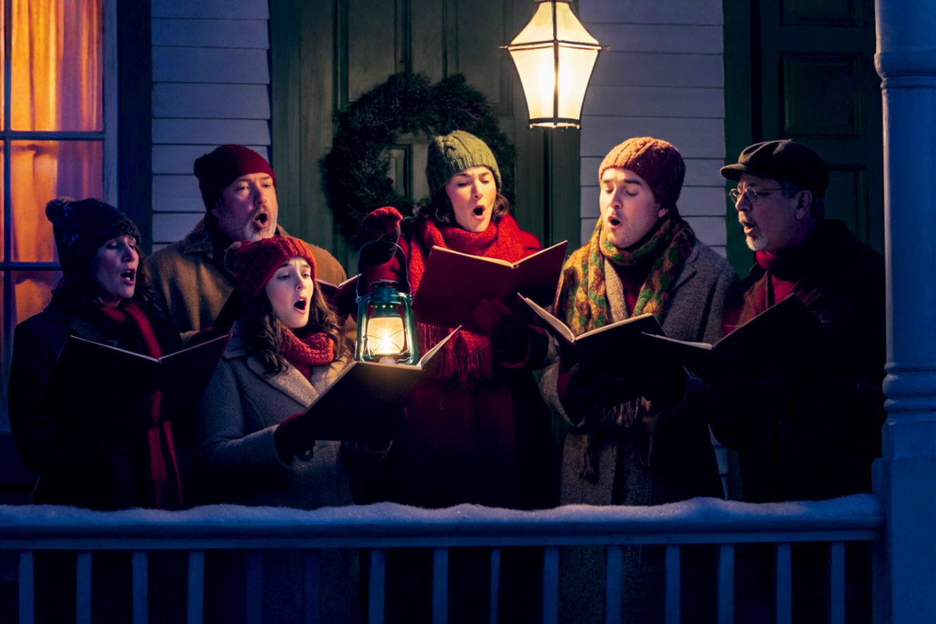 A professional, lifelike photograph capturing the warmth and magic of a small group of carolers on a snowy evening. The scene is set on the charming, lamplit porch of a historic home decorated for Christmas with a wreath on the door and glowing lights. The diverse group of four to five adults and children are bundled in cozy, classic winter attire (scarves, hats, wool coats) and are singing with joyful expressions, their breath visible in the cold air. One person holds a lantern that casts a warm, golden glow on their faces and the open songbooks they hold. The composition should be intimate and heartwarming, with soft, falling snow creating a magical atmosphere. The focus is on the singers' faces, capturing their genuine emotion and the spirit of community. Use a shallow depth of field to softly blur the background, emphasizing the carolers and the inviting glow of the home.