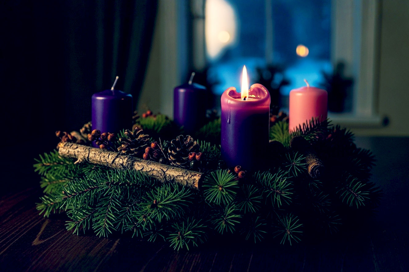 A professional, lifelike photograph of a rustic Advent wreath on a dark wooden table. One single purple candle is lit, its flame casting a warm, golden glow. Three other candles on the wreath—two purple and one pink—are not lit, their wicks dark and untouched. The wreath is made of fresh fir and pine branches. The camera is focused closely on the single flame, creating a shallow depth of field. The setting is a cozy, dimly lit room with soft, contemplative lighting. In the blurred background, a window looks out onto a snowy evening. The mood is peaceful, sacred, and full of hope.