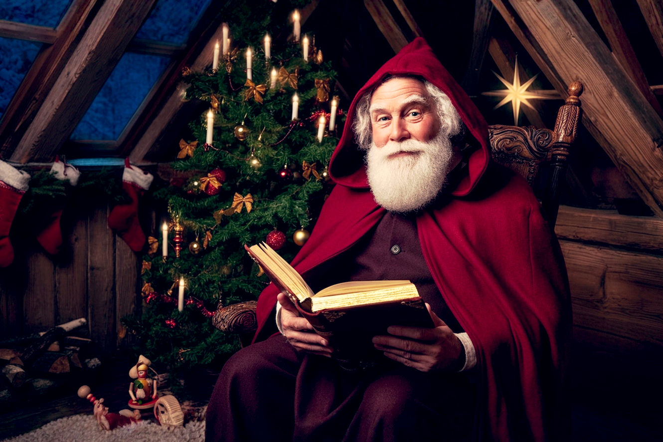 A highly detailed, professional photograph of a traditional German Weihnachtsmann in a cozy, dimly lit room with exposed wooden beams. He is a wise, kind-faced old man with a real white beard, wearing a deep red, hooded woolen cloak, not a bright modern suit. He is sitting next to a beautifully decorated Christmas tree, holding a large, leather-bound golden book on his lap and looking warmly at the camera. A small, handcrafted wooden toy sits on the floor beside him. The lighting is soft and warm, coming from the tree's candles and a nearby fireplace, creating a magical, nostalgic, and authentic atmosphere.