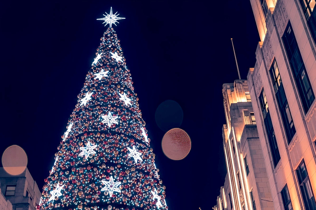 Professional photography of the Rockefeller Center Christmas Tree at night, illuminated with thousands of multi-colored LED lights, with the iconic Swarovski Star prominently displayed at the top. The composition should capture the tree's immense scale, with a slight upward angle emphasizing its height against the backdrop of the surrounding Art Deco buildings of Rockefeller Center. The image should be vibrant and sparkling, with a festive, magical, and slightly ethereal mood. Include soft bokeh lights in the background to enhance the holiday atmosphere. The style should be hyperrealistic and highly detailed, resembling a high-resolution, award-winning photograph.