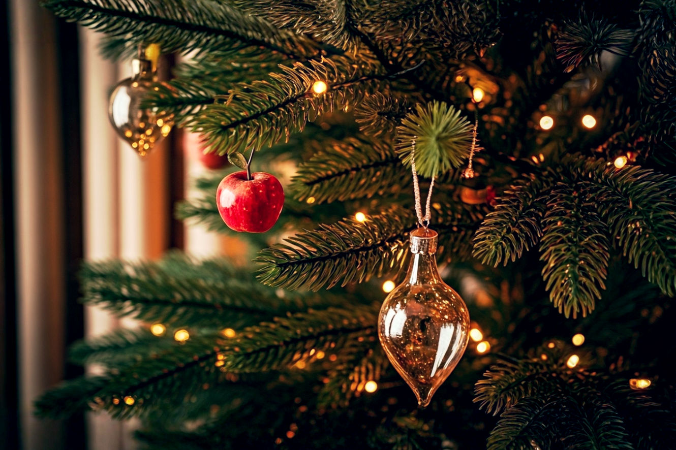 A stunning, high-resolution, lifelike professional photograph of a beautifully decorated, traditional Christmas tree, illuminated with warm, glowing lights. The tree should be adorned with a mix of historical and modern ornaments, subtly hinting at its rich origins: perhaps a few small, real red apples, some simple paper chains, a delicate glass Kugel, and strands of shimmering tinsel, alongside modern electric lights. The composition should be eye-level, focusing on the intricate details of the decorations, with a soft, blurred background of a cozy, festive living room. The lighting should be warm and inviting, emphasizing the glow of the lights and the sparkle of the ornaments, creating a mood of historical depth, warmth, and timeless holiday magic.